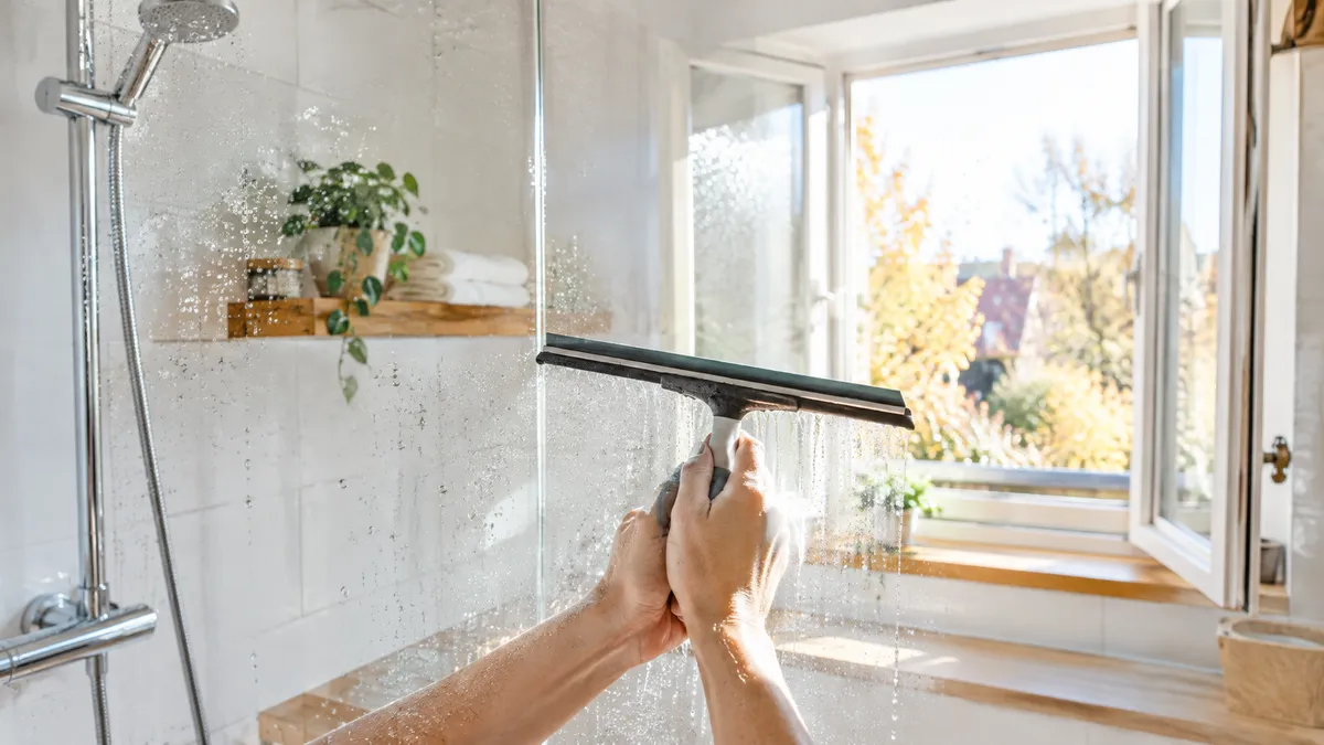 Hände ziehen nach dem Duschen einen Abzieher über eine Glasduschwand, im Hintergrund steht das Badfenster weit offen